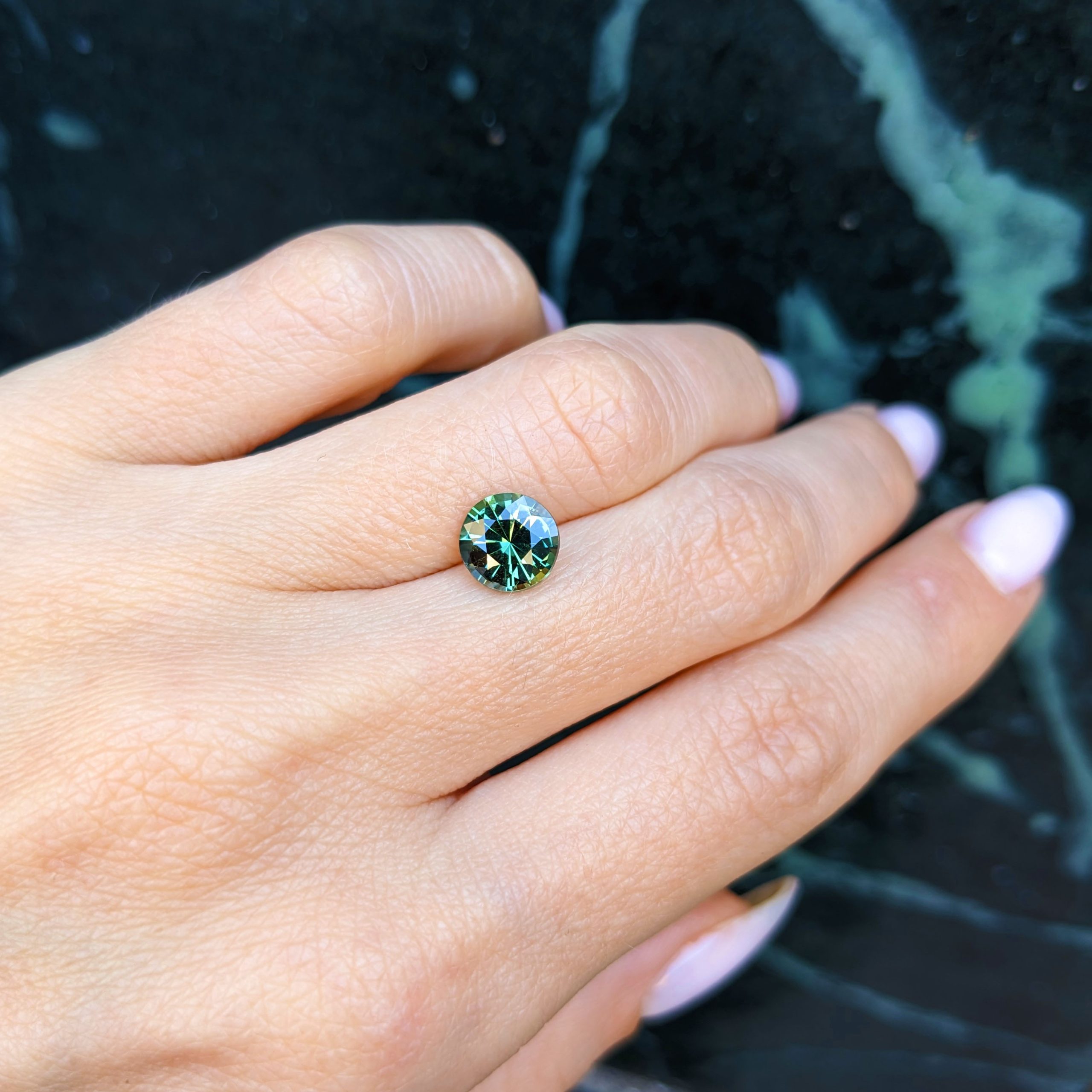 Natural 2.14 carat Madagascar bluish green round sapphire on a hand in front of dark marble